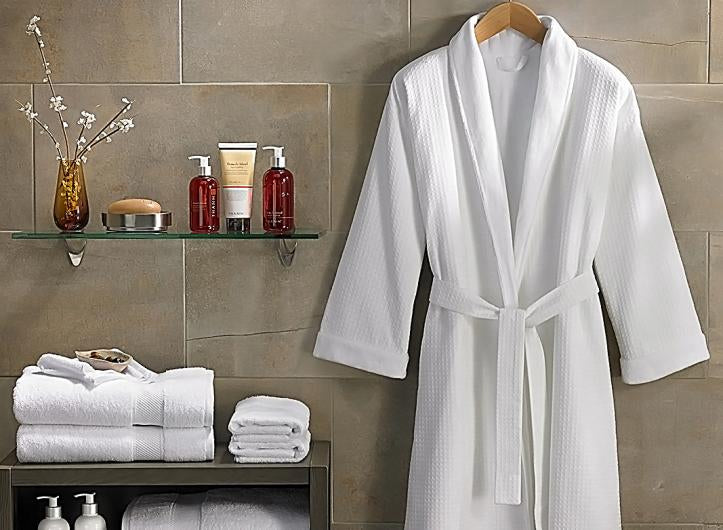 White bathrobe hanging on a wooden hanger next to folded towels and bathroom products on a shelf.